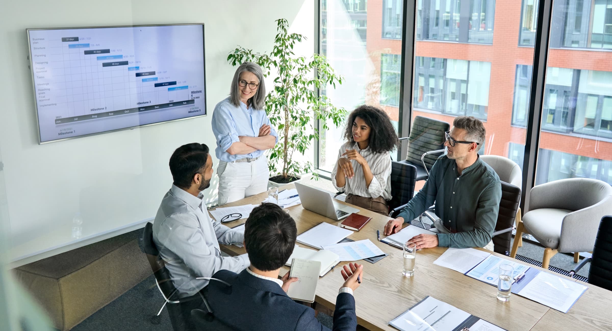 A leader presenting to a seated executive team in a boardroom, with a project roadmap on the wall screen.